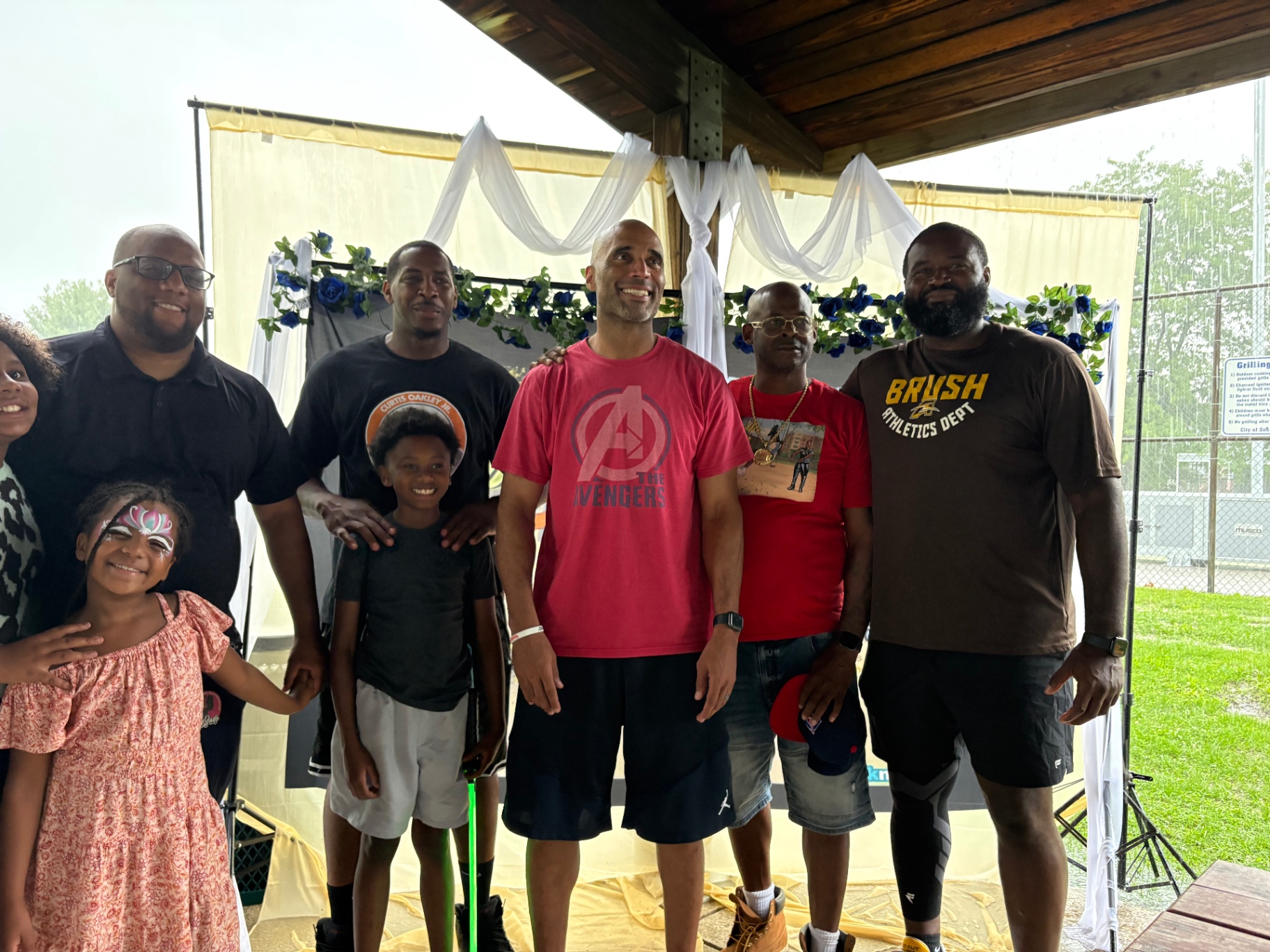 A group of Black fathers, a young boy and two girls smiling in front of the blue-flowered Push Me Dad backdrop