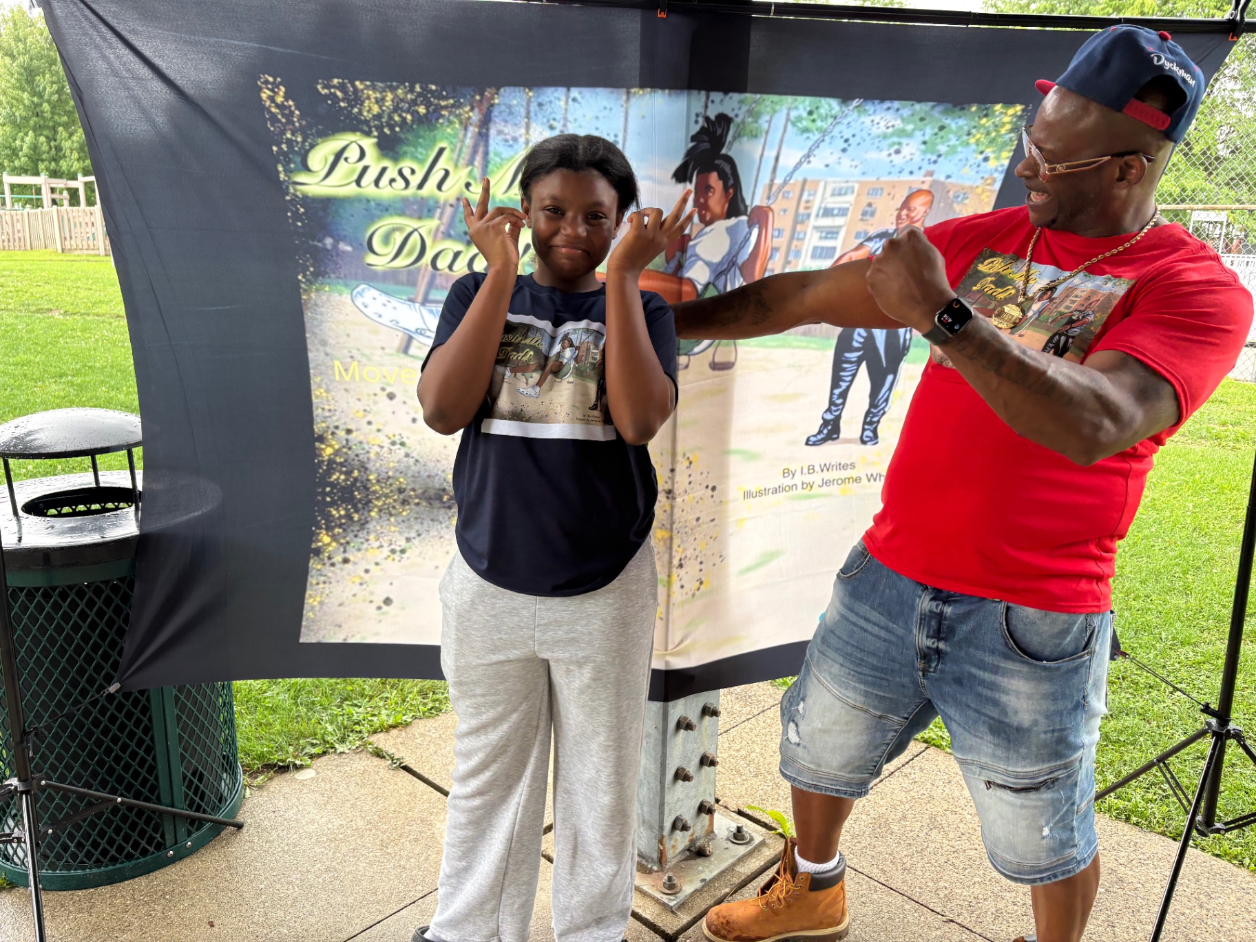 The Push Me Dad founder and his daughter laughing and playing in front of the book banner at a park celebration