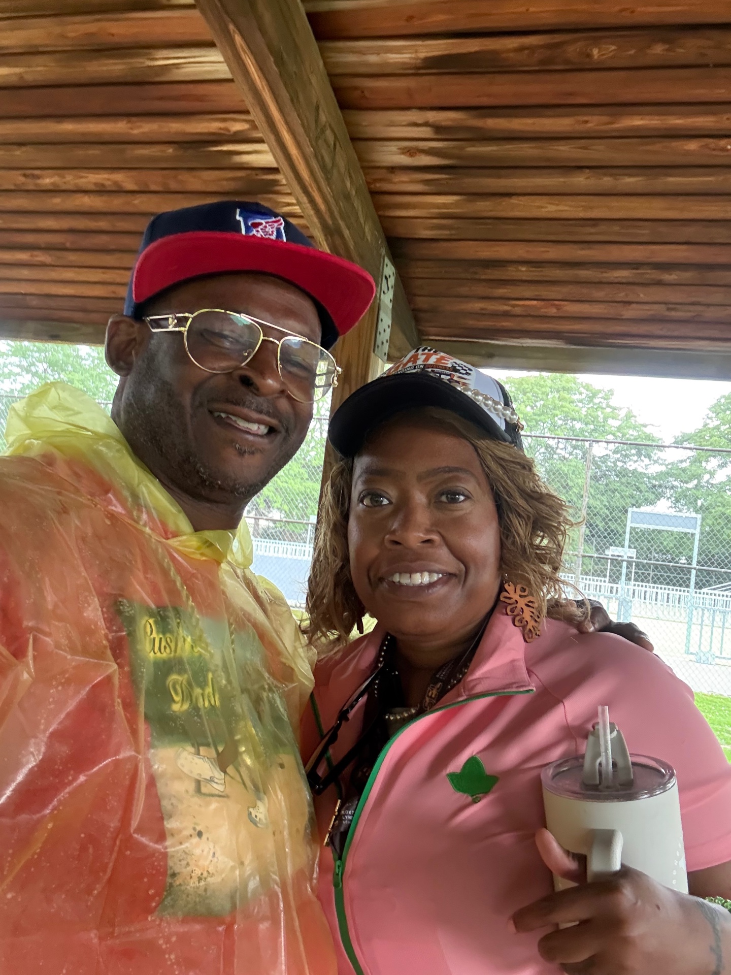 A father in a Push Me Dad shirt and rain poncho smiling with a supporter under the pavilion during a rain shower