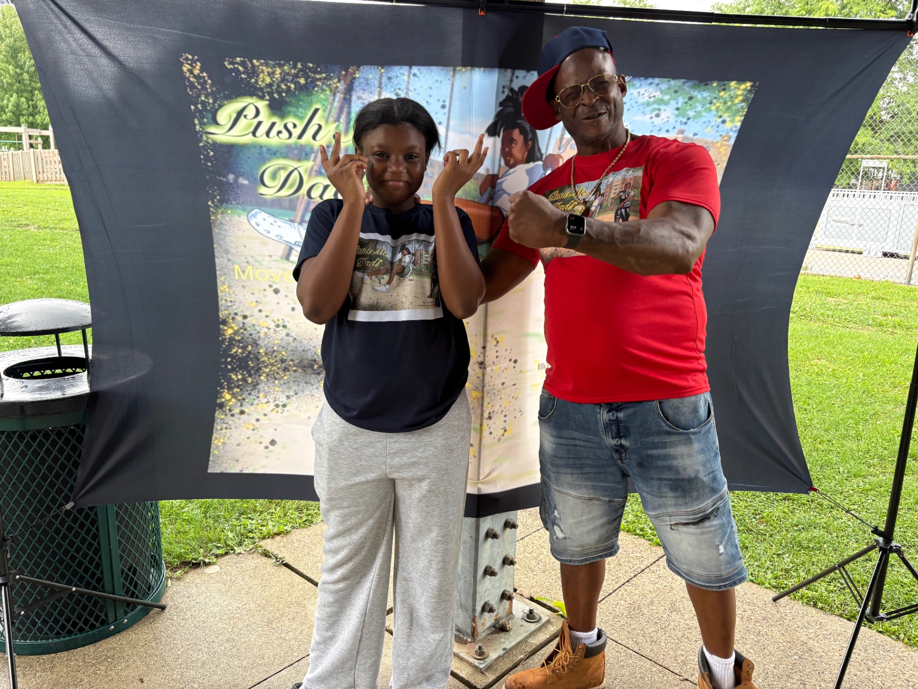 The founder flexing alongside his smiling daughter in front of the Push Me Dad book backdrop at Bexley Park