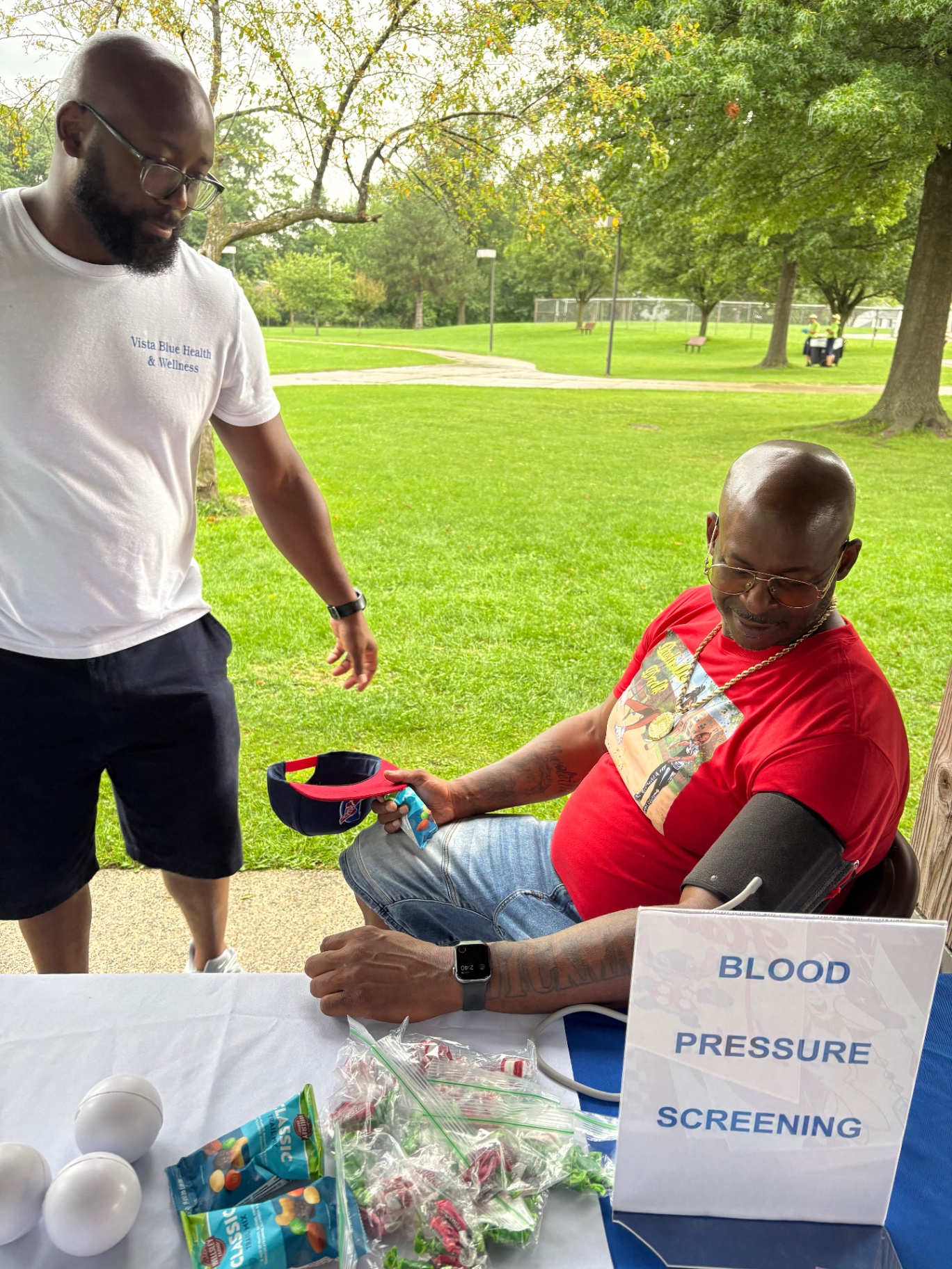A Vista Blue Health & Wellness volunteer talking with a seated father wearing a blood pressure cuff at the free screening table