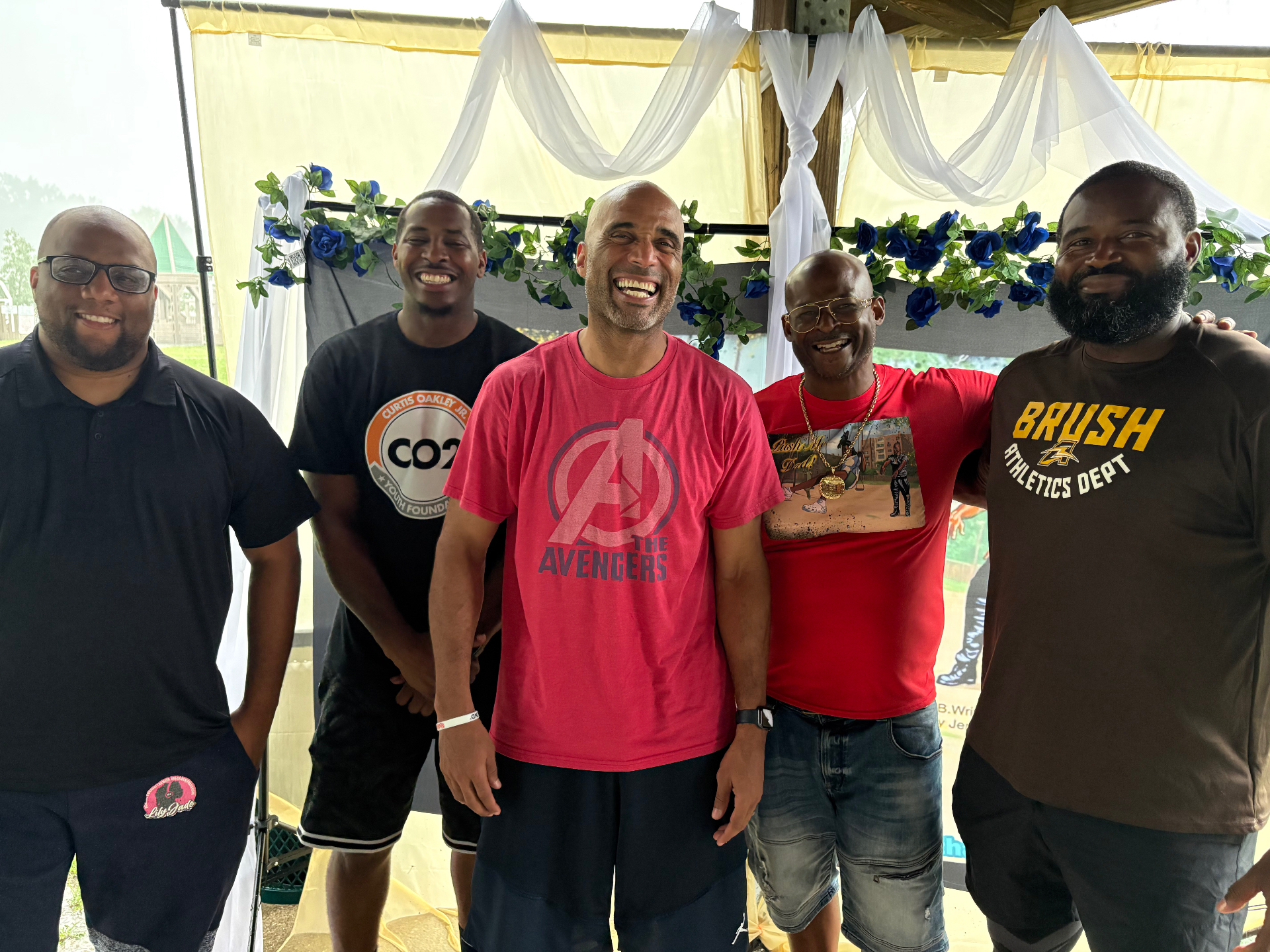 Five Black fathers laughing together arm in arm in front of the blue-rose Push Me Dad backdrop under the pavilion