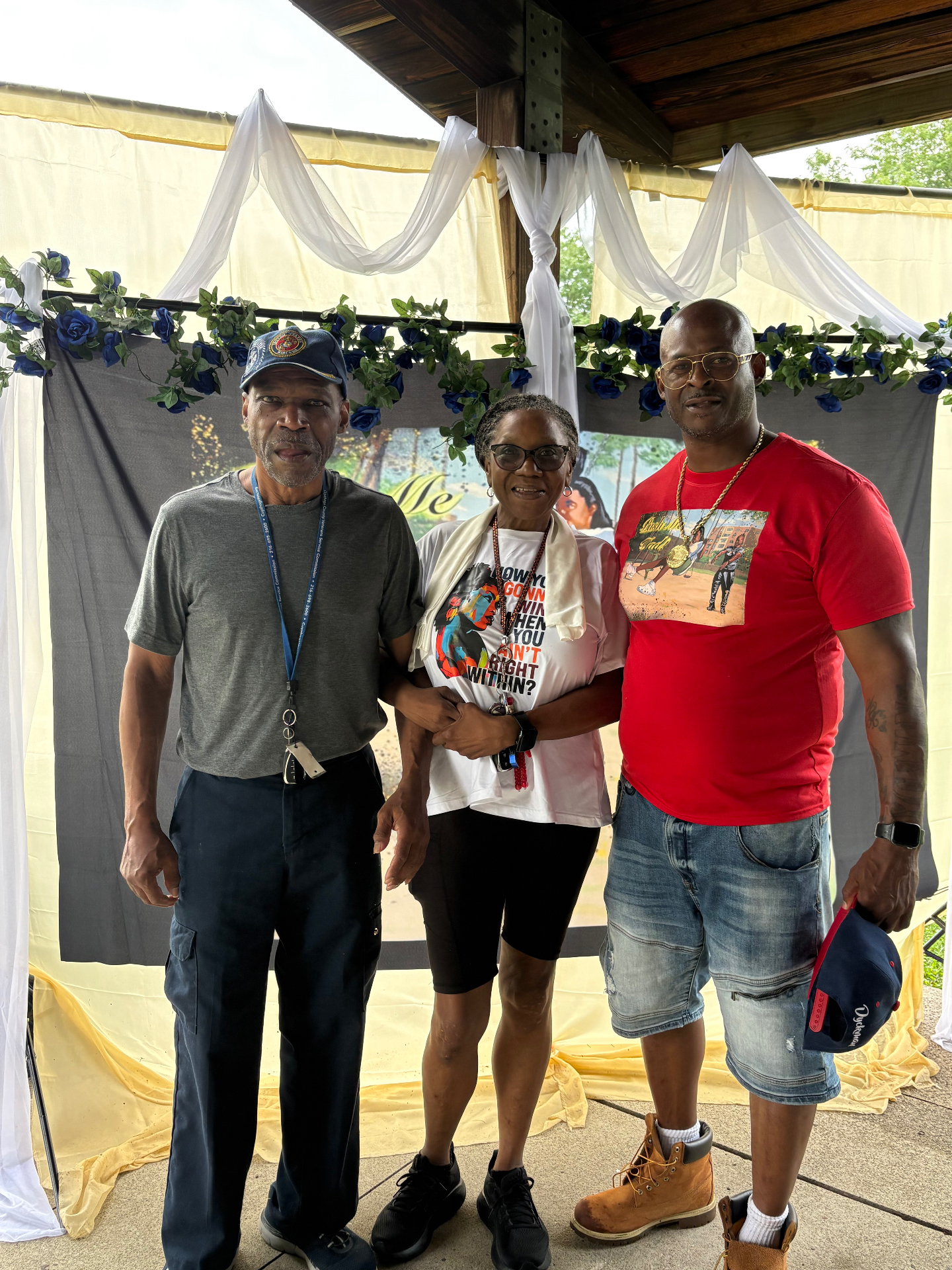 Two Black men and a woman standing arm in arm in front of the blue-rose Push Me Dad backdrop under the pavilion