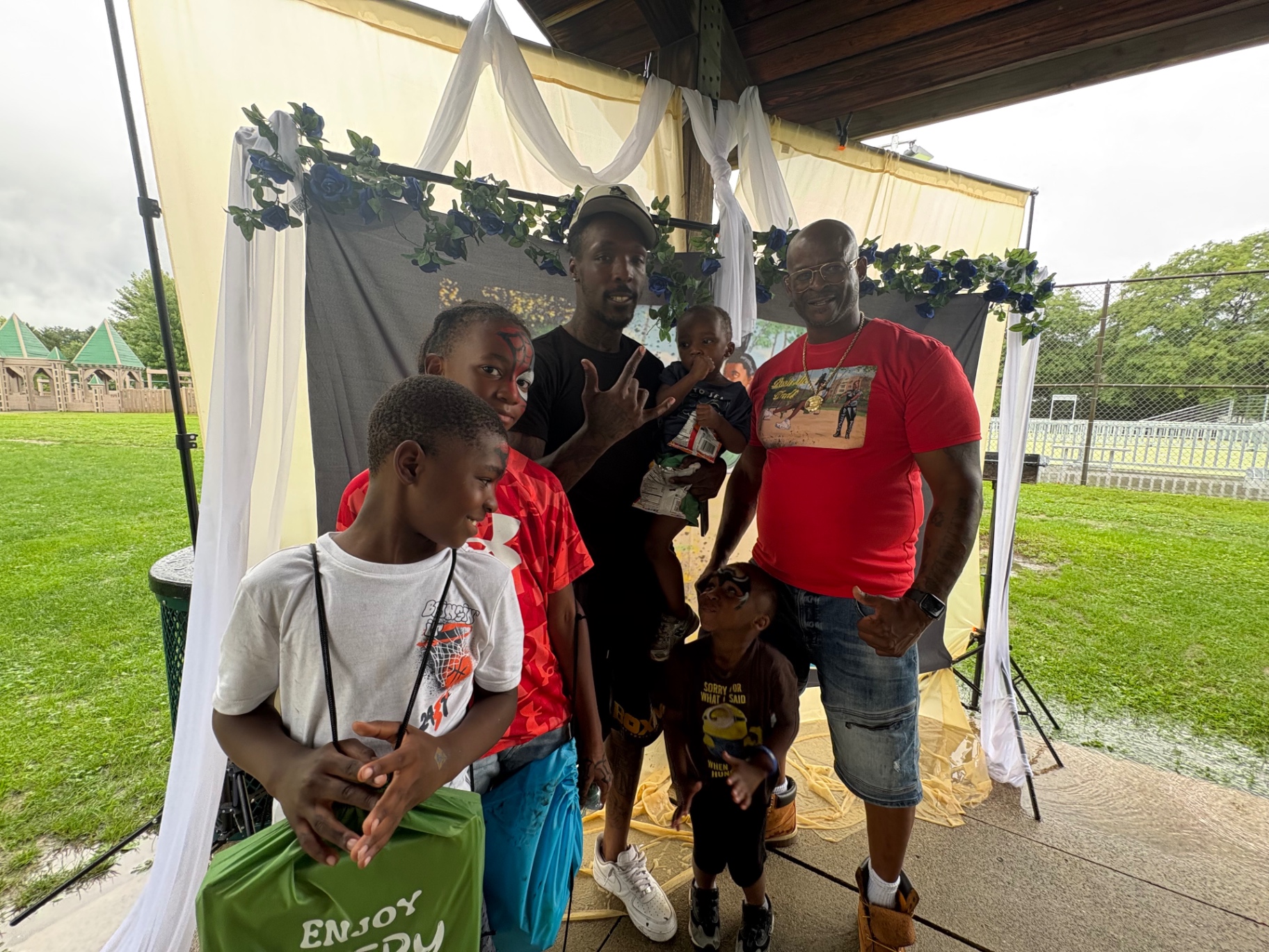 A Black father holding his toddler while surrounded by children with face paint at a Push Me Dad park celebration