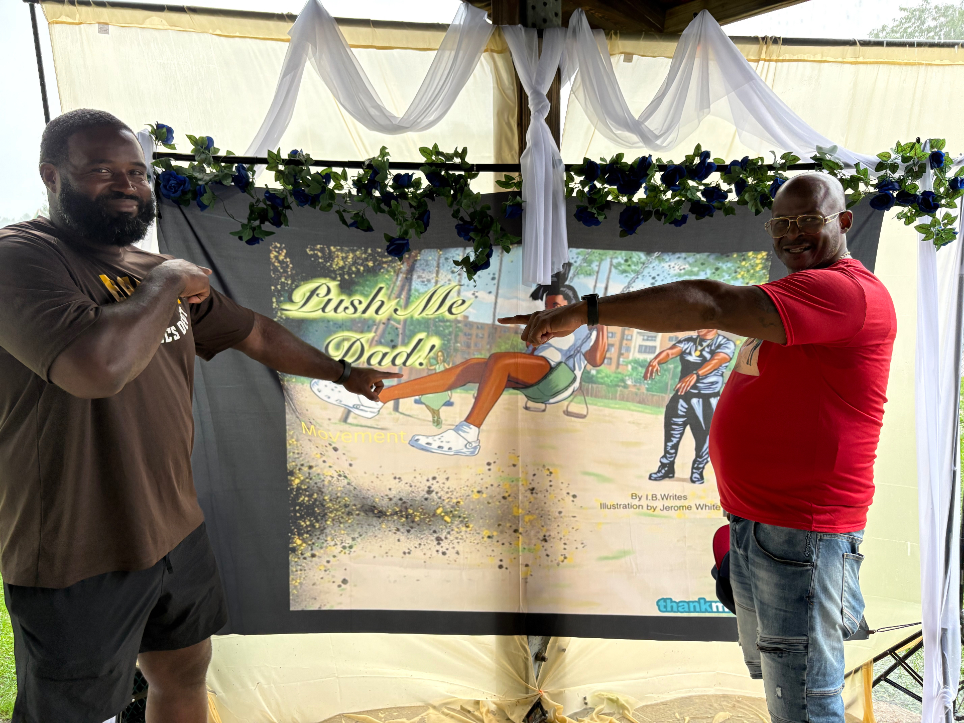 Two Black fathers smiling and pointing toward the Push Me Dad book banner at Bexley Park