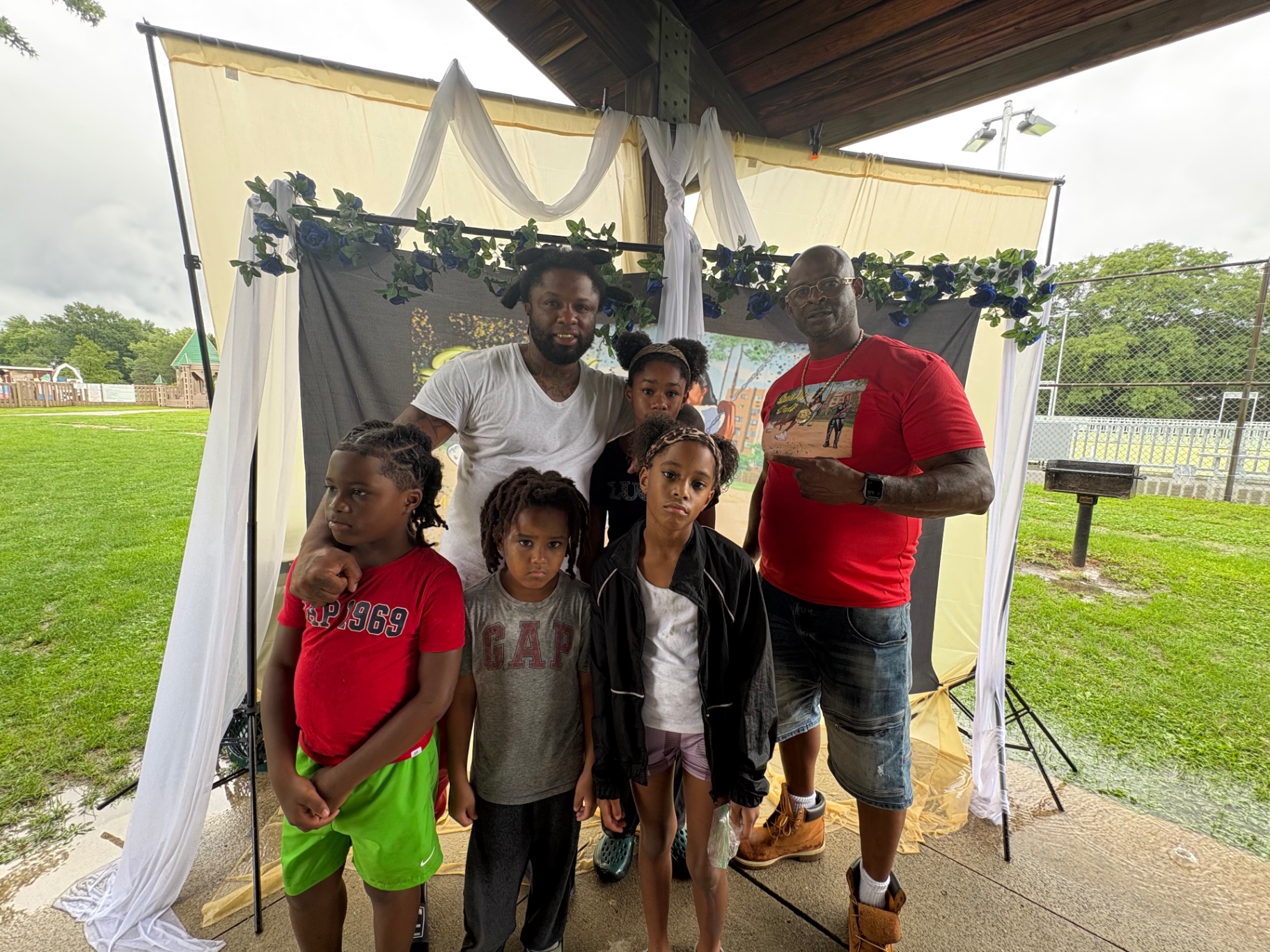 Two Black fathers standing with five children in front of the decorated Push Me Dad backdrop at a park event