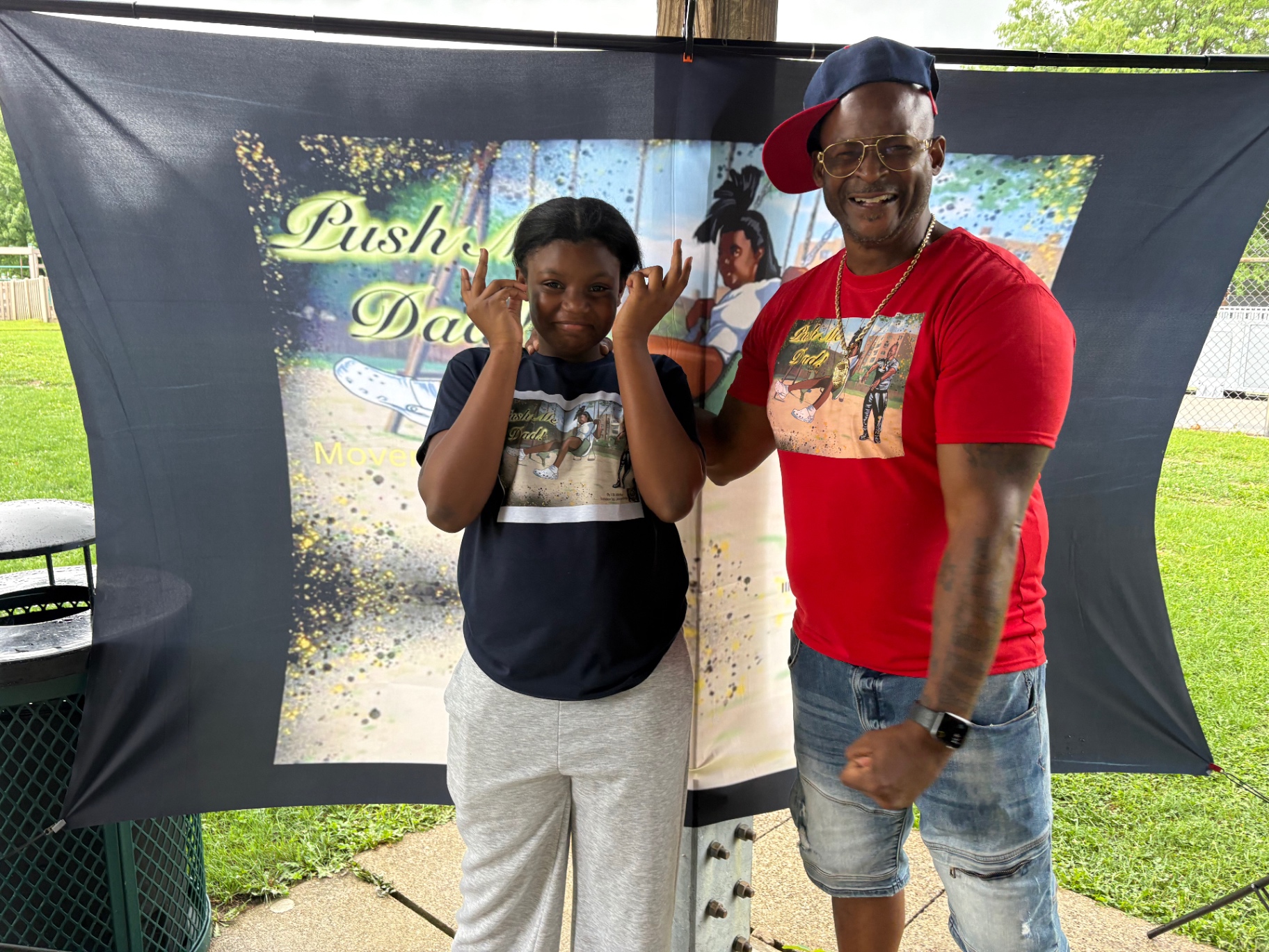 Push Me Dad founder in a red Push Me Dad t-shirt posing with his daughter in front of the Push Me Dad book banner at a community park event