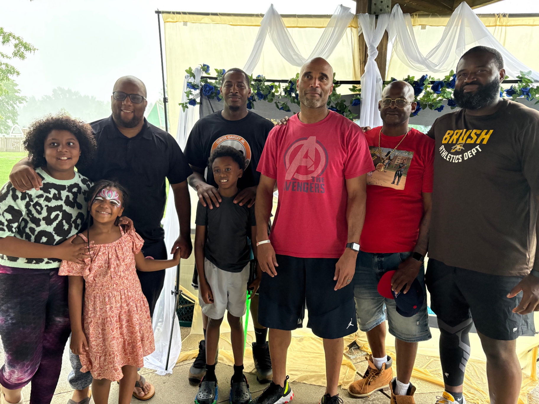Five Black fathers and four children posing together in front of the Push Me Dad banner under the pavilion at Bexley Park