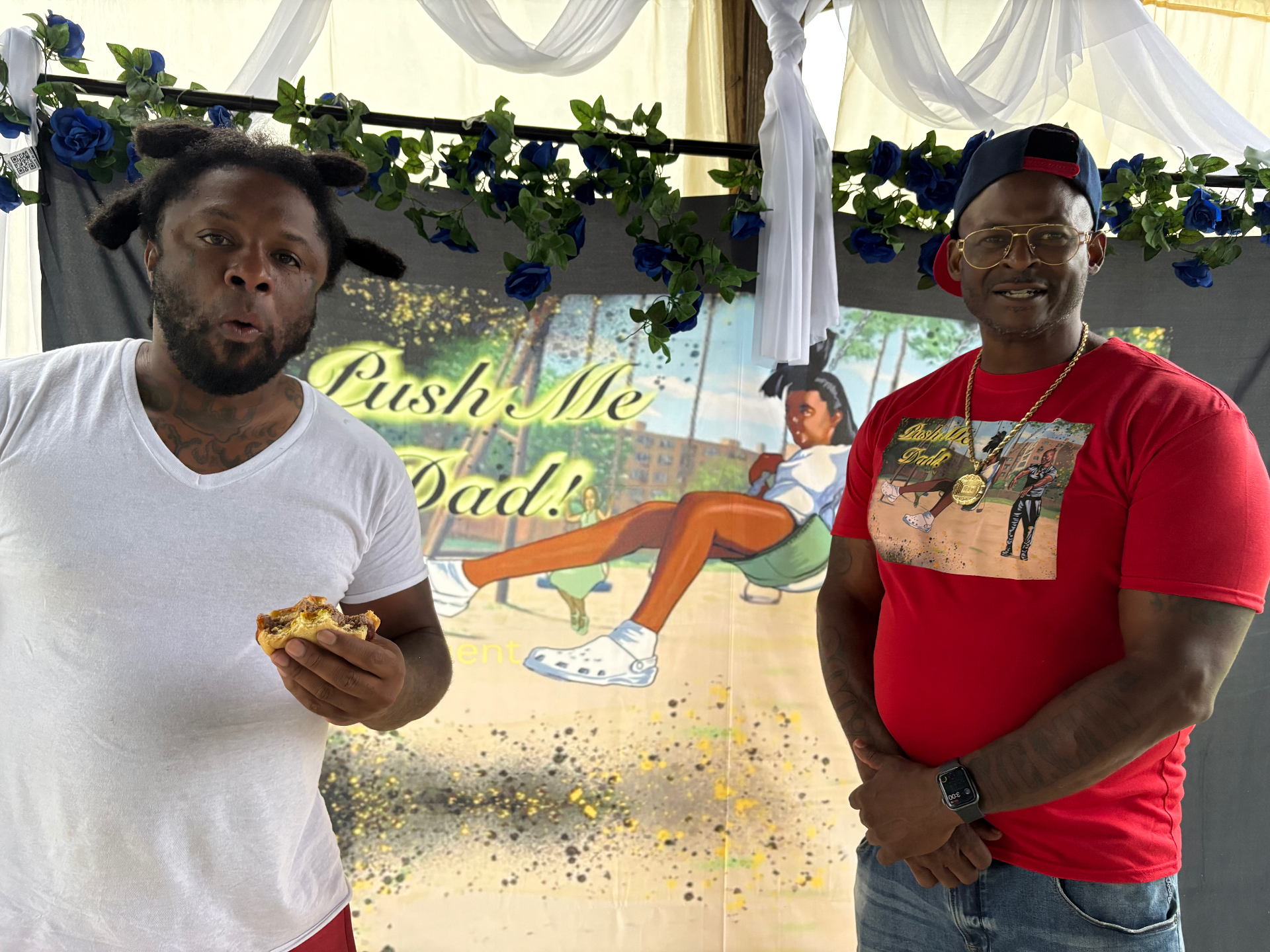 Two Black men, one holding a burger, standing in front of the Push Me Dad book banner at the cookout
