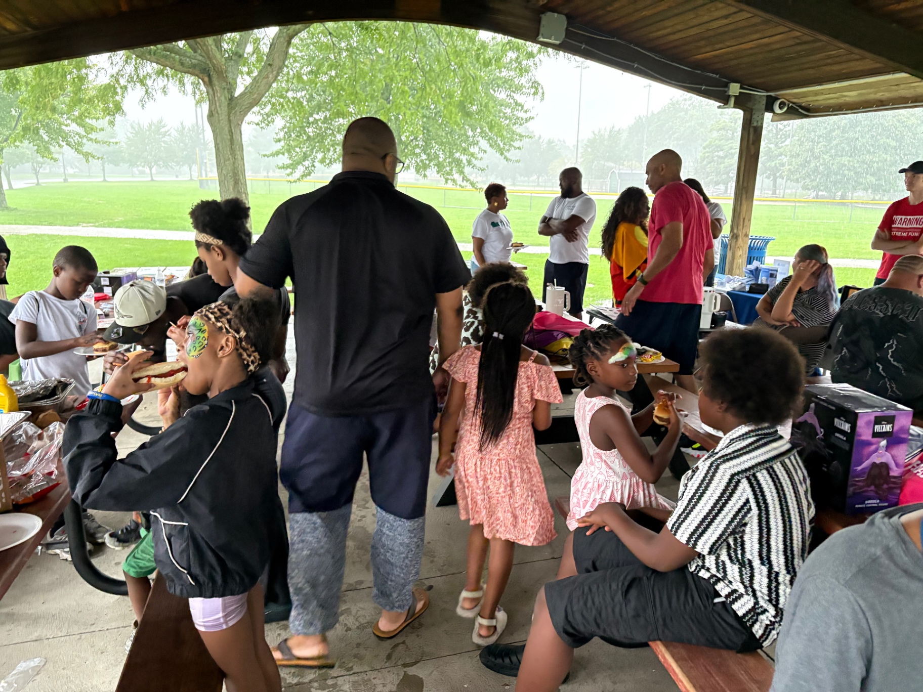Families eating burgers and hot dogs at picnic tables under the pavilion during the Push Me Dad kickball celebration